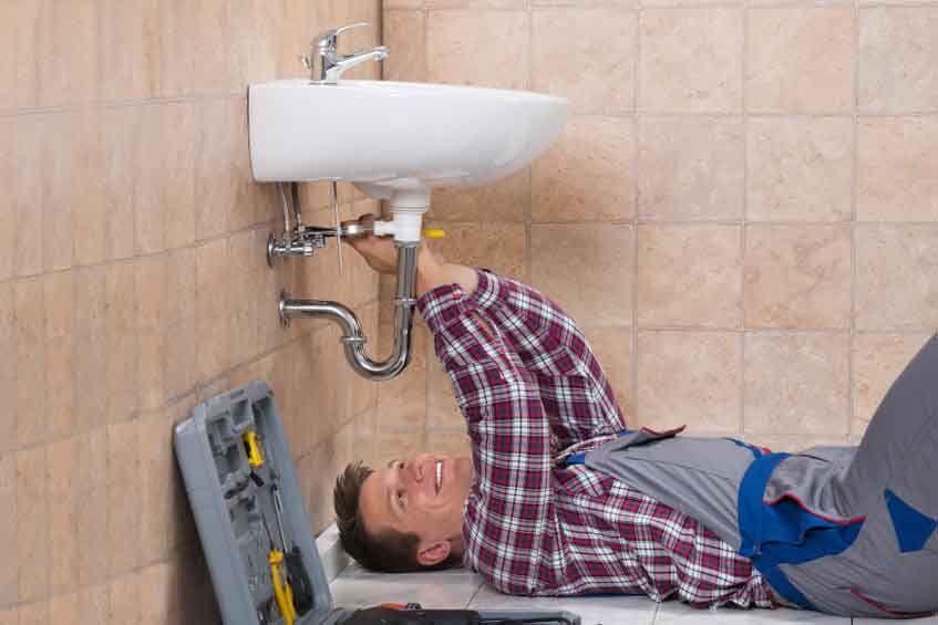 Plumber Lying On Floor Fixing Sink Happy Young Male Plumber Lying On Floor Fixing Sink In Bathroom